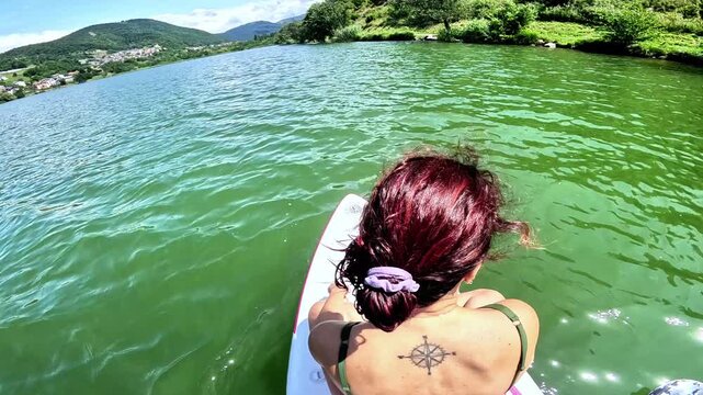 Slow-motion of a woman sitting on a stand-up paddle board in a lake surrounded by hills framed from behind with upward chamber movement. Lake Serraia is located in provence of Trento, in Trentino