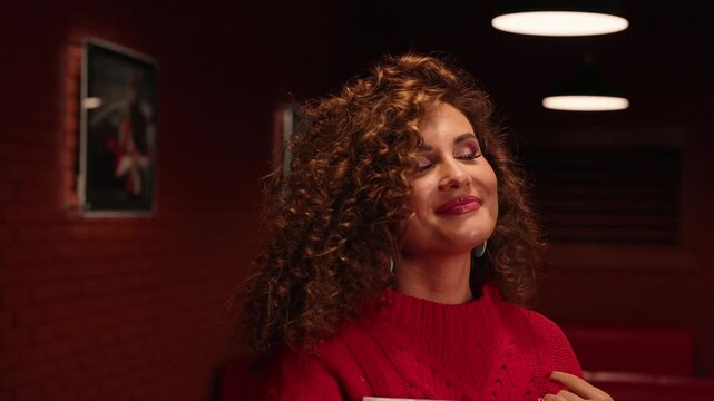 Dim red room with brick walls and vintage photos. Soft lighting creates warm ambiance. She smiles gently while holding a book. Close-up reveals relaxed, inviting expression