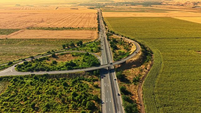 A road connects across fields of green and yellow. The scene shows farms and crops on both sides. Cars travel along the road, and the weather is sunny.