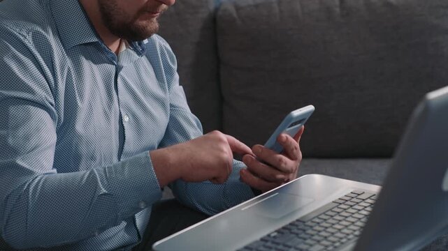 Man using smartphone next to laptop during work switching between devices and completing online tasks in calm home environment