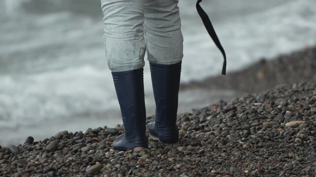 Rubber boots walking on wet pebble beach beside crashing ocean waves during rough weather exploration.