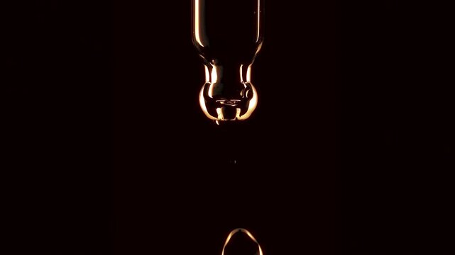 Macro shot of a golden vitamin C ascorbic acid or argan oil droplet falling from a glass pipette dropper in slow motion isolated on a dark background
