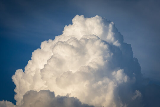 Towering storm built clouds reflect in a lake like a tall mirror while a wooded hill frames the scene creating a dramatic peaceful view of weather and landscape in late day light