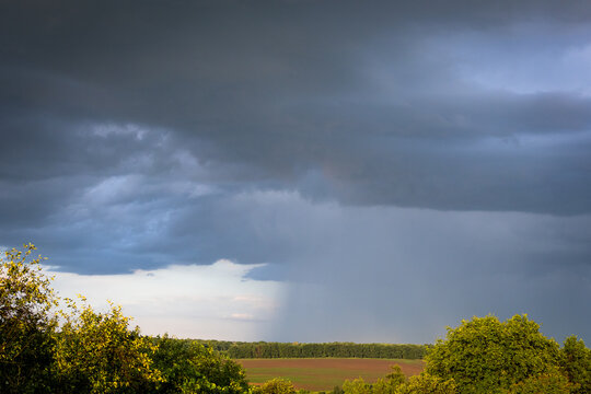 A storm sweeps across open farmland with a visible rain core beneath towering clouds while patches of light touch the treeline capturing the restless energy of changing summer weather
