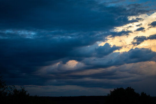 Brooding clouds gather over a quiet horizon with pale light breaking through layered textures suggesting passing showers and unsettled conditions during a tranquil summer twilight