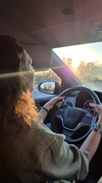 Woman driving car during golden hour as she navigates urban streets while sun glares. Handheld shot with side profile view.