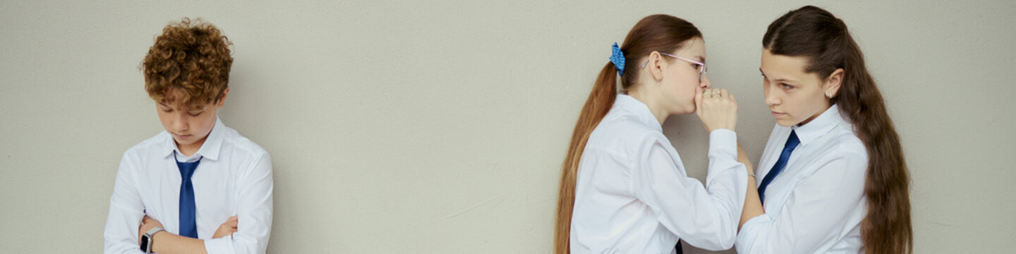 Header of teenage schoolgirls whispering while teenage boy standing apart with crossed arms, showing exclusion and bullying. Useful for school conflict, peer pressure