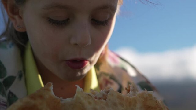 Girl, eating, outdoors. Young child bites heartily into large street food bread against a bright blue sky on a sunny summer day.