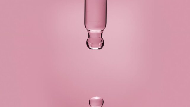 Macro shot of a rose water extract or vitamin B12 serum droplet falling from a glass pipette dropper in slow motion isolated on a solid pink background