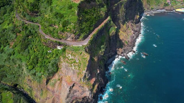 Ponta Delgada North Madeira drone down to top view of old cliffside road in green mountains, coastal village on protruding peninsula and dramatic Atlantic ocean