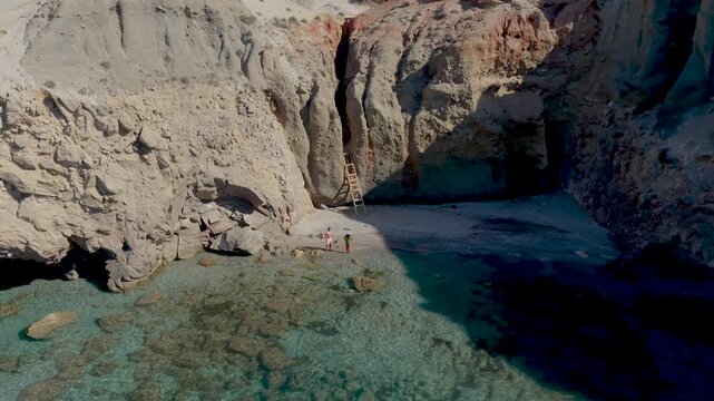 A couple of men and women walk at the Tsigrado Beach Milos, Greece, where turquoise waters meet majestic cliffs. Enjoy a serene atmosphere while relaxing on the sandy shores of this stunning island