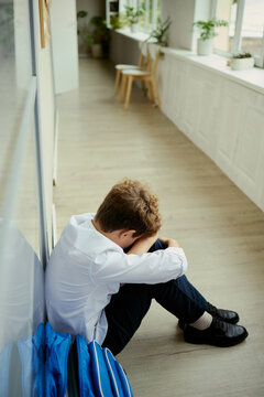 Vertical shot of Caucasian schoolboy sitting on floor in hallway, hiding face in arms, expressing school stress, loneliness, bullying concept. For education, child mental health campaigns