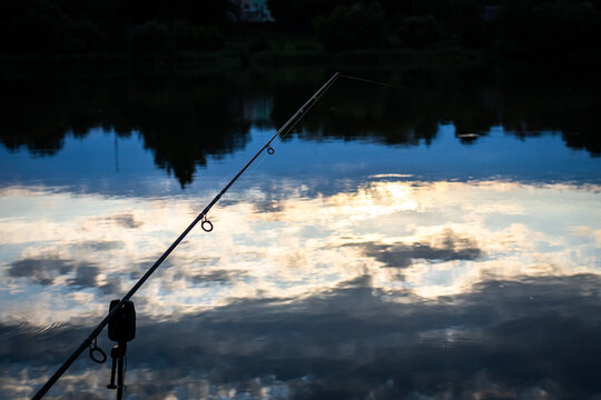 A single rod stands above cool blue reflections in the last light of day. The bite alarm rests silent while clouds fade and the lake becomes a mirror inviting slow breathing and focus.