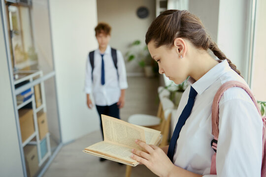 Teenage girl reading book while standing indoors with male classmate in background, showing study focus and school routine. Useful for education campaigns, teen learning content