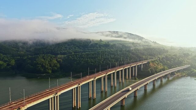 Long elevated bridge carries moving vehicles across calm river while low fog drifts over forested hills, creating peaceful dawn scenery with strong sense of travel, distance, and scale