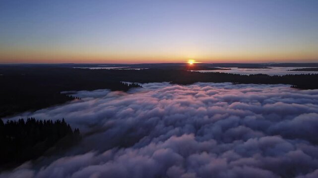 Aerial view of dense fog covering forested landscape at dawn, transitioning to sunrise illuminating the horizon over a serene lake and rolling hills