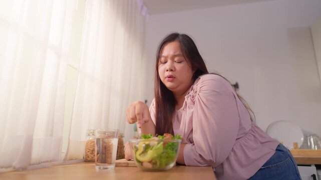 Asian plus-size woman looking unhappy and reluctant to eat green salad. 