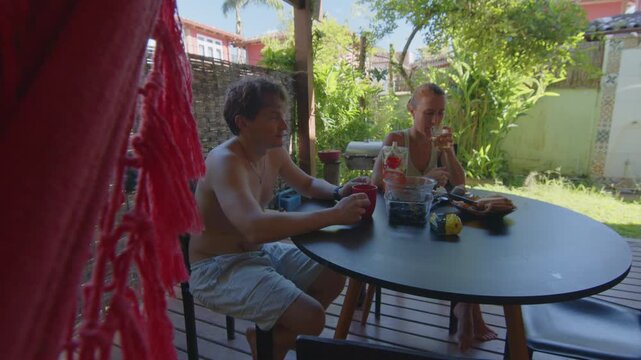 Two people having breakfast and talking at an outdoor patio table in a tropical garden