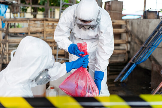 Hazardous material workers in protective clothing lifting a red toxic waste bag behind yellow caution tape during a chemical spill decontamination process to maintain industrial hygiene.