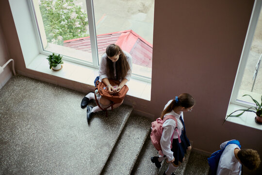 High angle view of school teenagers moving through stairwell while girl sitting by window checking smartphone, showing student routine and distraction. Useful for education, youth behavior campaigns