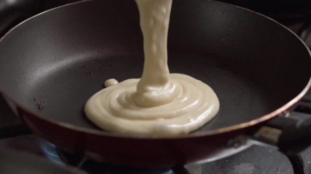 Fresh pancake batter pouring into hot frying pan over gas flame during breakfast preparation. Home cooking process showing smooth mixture spreading before flipping stage