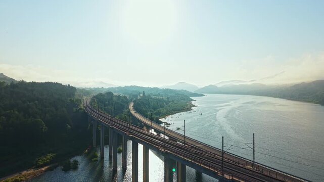 Long freight train moves across elevated concrete and steel bridge above calm reflective river, showing forward motion toward distant shoreline with repeating piers, open water and aerial landscape