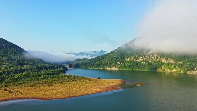 Elevated view of broad river bend with gentle flowing water, subtle surface ripples, drifting mist, forested hills, sandy pebble spit, wet shoreline, exposed rocks, soft daylight, and peaceful