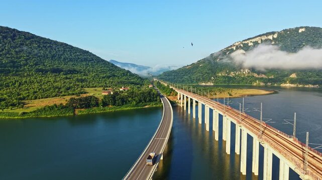 Long concrete bridge carries vehicles across wide river between green hills while traffic moves steadily through soft mist and warm sunlight, with calm reflections below, with bridge, highway, river
