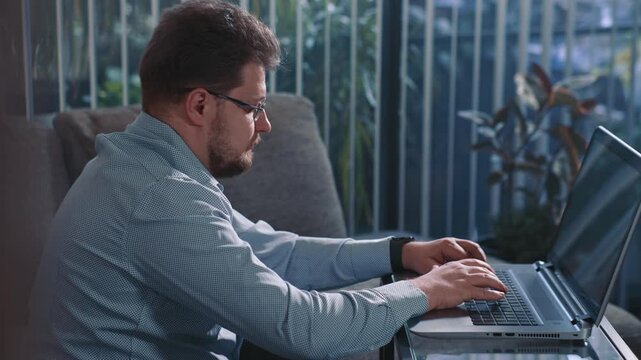 Man working on laptop at home sitting near window typing on keyboard and completing online tasks of remote work in calm home environment