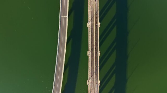 Top-down aerial scene showing two parallel bridge spans crossing deep green water, modern concrete roadway with lane markings and moving vehicles beside rust-toned steel truss, creating striking