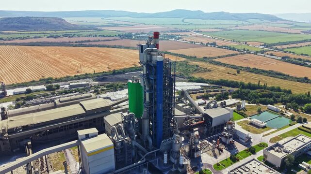 A large cement factory is seen from above, set against open fields and mountains. The factory has tall structures and colorful parts. It is a clear sunny day.