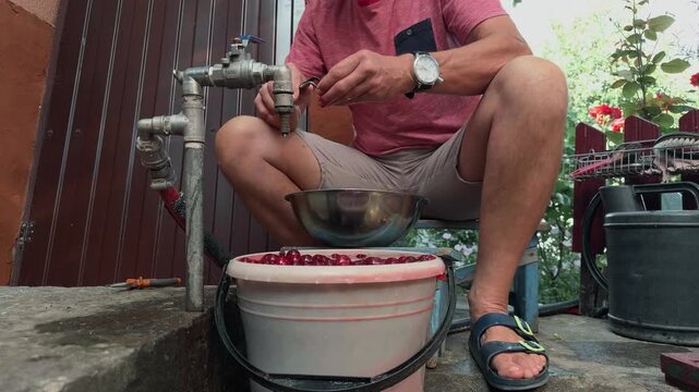Unrecognizable adult man pitting fresh red cherries with metal tool outdoors in summer garden sitting by water tap low angle authentic rural domestic lifestyle handheld shot of fruit preparation