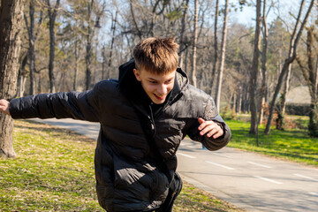 Fototapeta premium A teenage boy leans forward with arms spread wide in a playful dynamic pose on a sunny park path, with tall bare trees and green grass stretching behind him