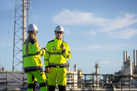 Professional workers in safety gear and high visibility jackets at petrochemical facility, male and female engineers with walkie talkie discussing work at oil refinery plant