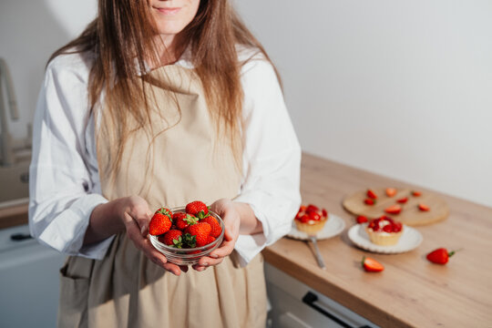Woman holding bowl of strawberries near berry tartlets on kitchen table