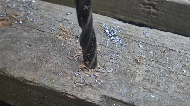Metal drill bit bores through a steel angle bracket piece during workshop fabrication. Extreme close-up shot.