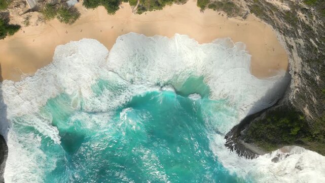 Aerial top-down view of turquoise waves crashing onto the golden sand of Kelingking Beach, Nusa Penida, Bali, Indonesia. Tropical coastline with dramatic cliffs, clear water, and iconic island scenery