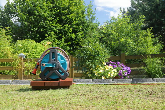 A garden hose reel with a sprayer nozzle on it stands on a wooden pedestal in front of a yellow wooden hedge in a summer garden