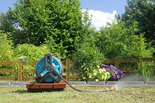 A garden hose reel with a sprayer nozzle on it is standing on a wooden pedestal and spraying water on the plants in front of a yellow wooden hedge in a summer garden.