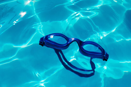 Swimming goggles on blue water surface in summer swimming pool with sunlight caustic. Underwater spectacles for diving, leisure and vacation.