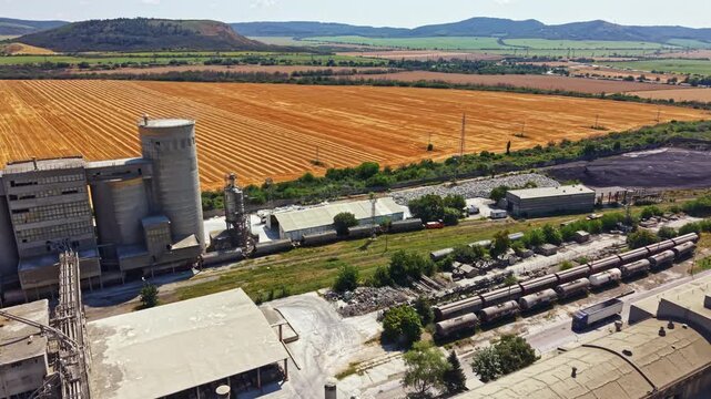 Large buildings and equipment are seen near train cars and roads. Fields of crops stretch out under the clear sky. Hills fade into the distance and the sun shines brightly on the scene.