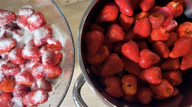 Fresh strawberries in a metal pot beside sugared strawberries in a glass bowl, top view kitchen prep scene, homemade jam making, summer fruit dessert ingredients