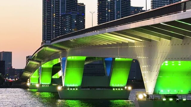 夕暮れのマジックアワーに鮮やかな緑と青のライトアップが映える橋脚構造と、背景にそびえる高層タワーマンション群。