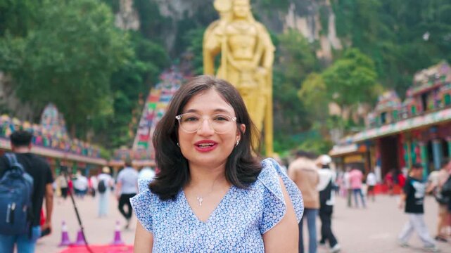 Portrait of young Indian woman vlogging while standing against famous Batu cave in Kuala Lumpur, Malaysia. Confident travel vlogger sharing travel experiences. Statue of Lord Murugan in background.