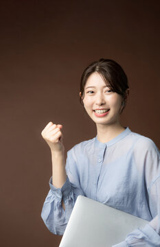  茶色背景でノートパソコンを持ってガッツポーズをする笑顔の日本人女性　仕事の成功・目標達成・応援の秋イメージ