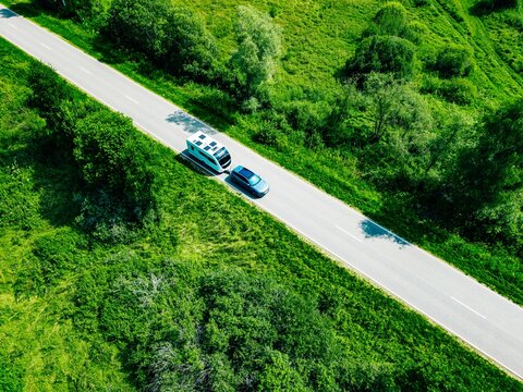 Aerial view of a blue modern car towing a caravan on a rural road through summer fields
