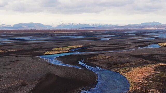 Sweeping aerial footage of a vivid blue glacial river winding through a vast black sand outwash plain in Iceland. The expansive volcanic landscape is framed by distant snow-capped mountains under a dy