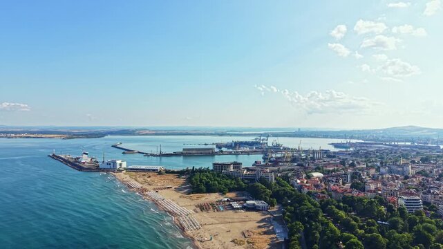 A coastal city stretches along the shore with sandy beaches and green trees. Ships are docked in the harbor while buildings line the waterfront. People enjoy the sun.