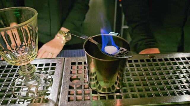 Close-up of bartender setting down an empty goblet beside blue flames, sugar cube, slotted spoon, metal cup, stainless bar counter, and moody cocktail lounge lighting indoors at night.