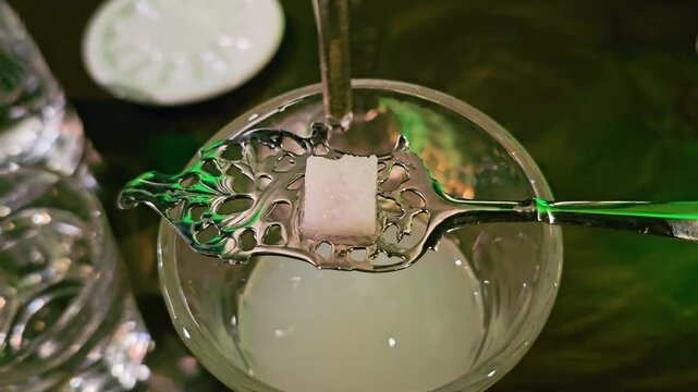 Absinthe louche ritual in a bar, with water droplet falling onto a sugar cube on a decorative spoon, cloudy drink below, vintage glassware, and atmospheric green lounge light.